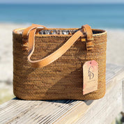 A woven straw handbag with a tag attached, placed on a wooden surface near the ocean.