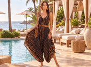 A woman in a black and gold patterned jumpsuit stands near a pool, with palm trees and a beach visible in the background.