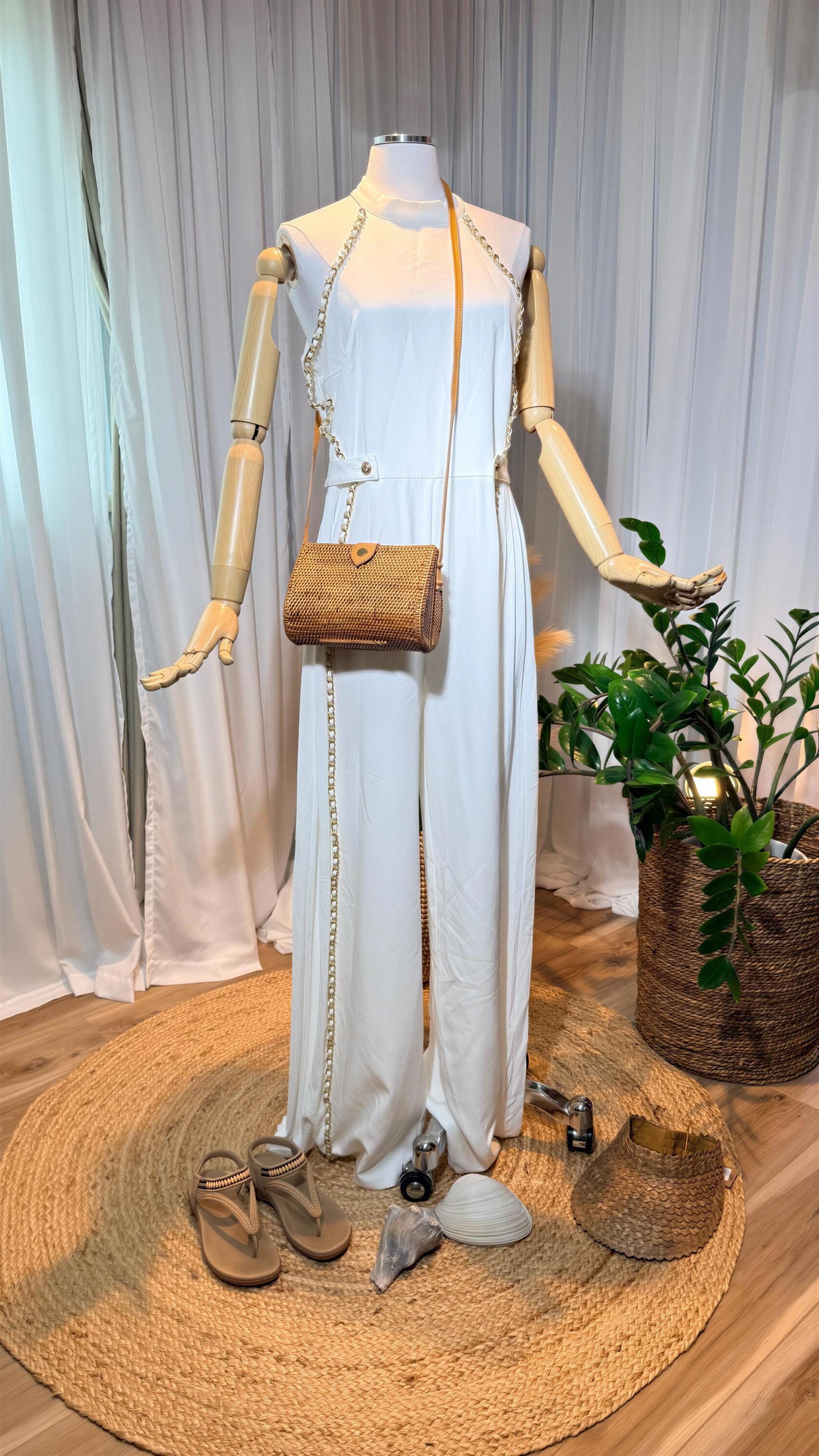 A mannequin wearing a white jumpsuit with a woven purse and sandals is displayed in a room with a woven mat and a potted plant.