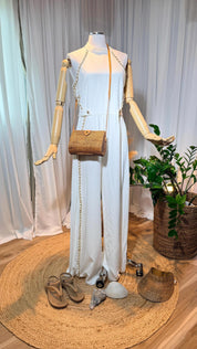 A mannequin wearing a white jumpsuit with a woven purse and sandals is displayed in a room with a woven mat and a potted plant.