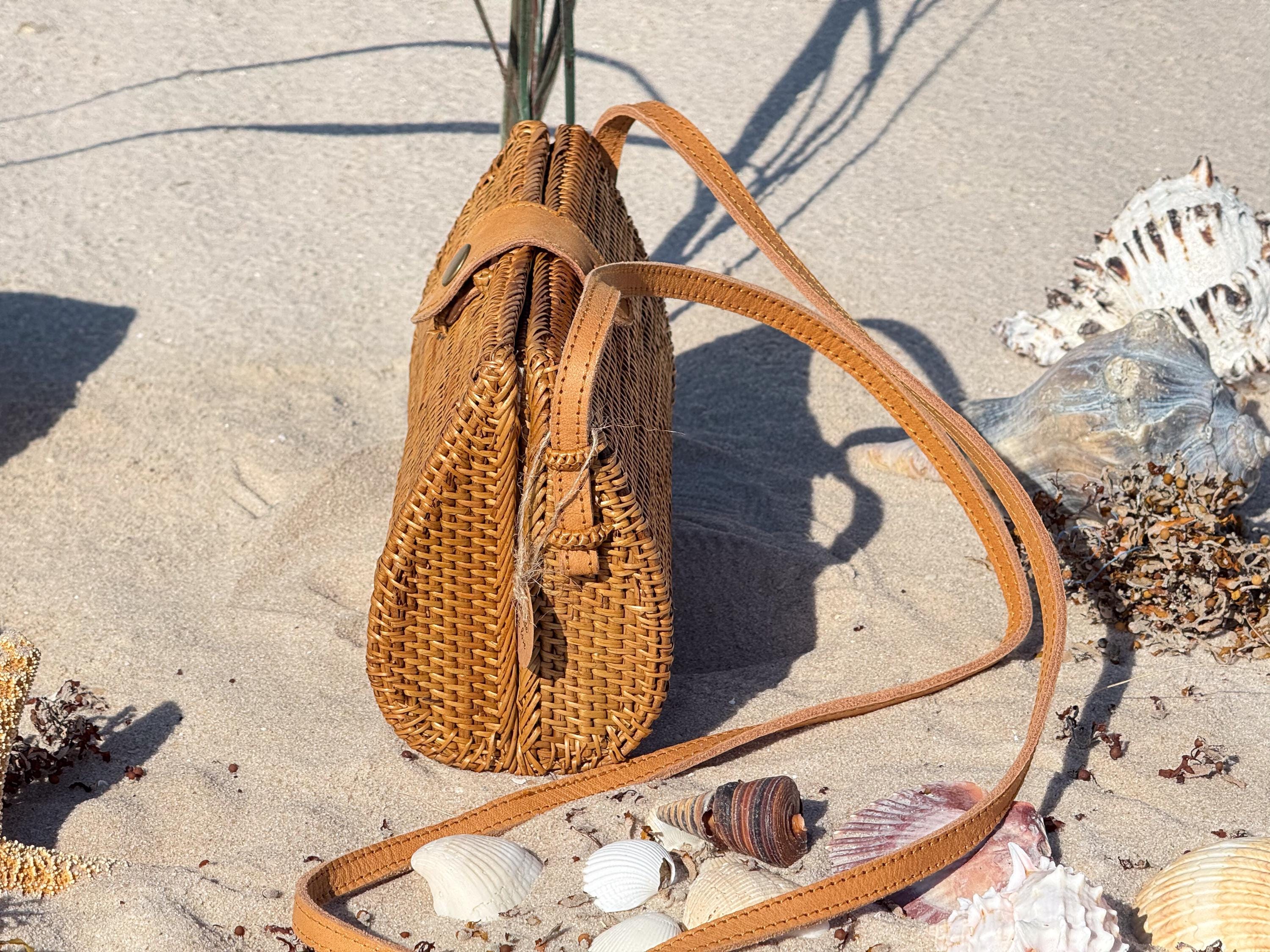 A brown woven purse rests on a sandy beach, surrounded by seashells and other beach items.