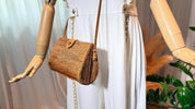 A mannequin wearing a white jumpsuit and carrying a brown woven purse is displayed in a store window.