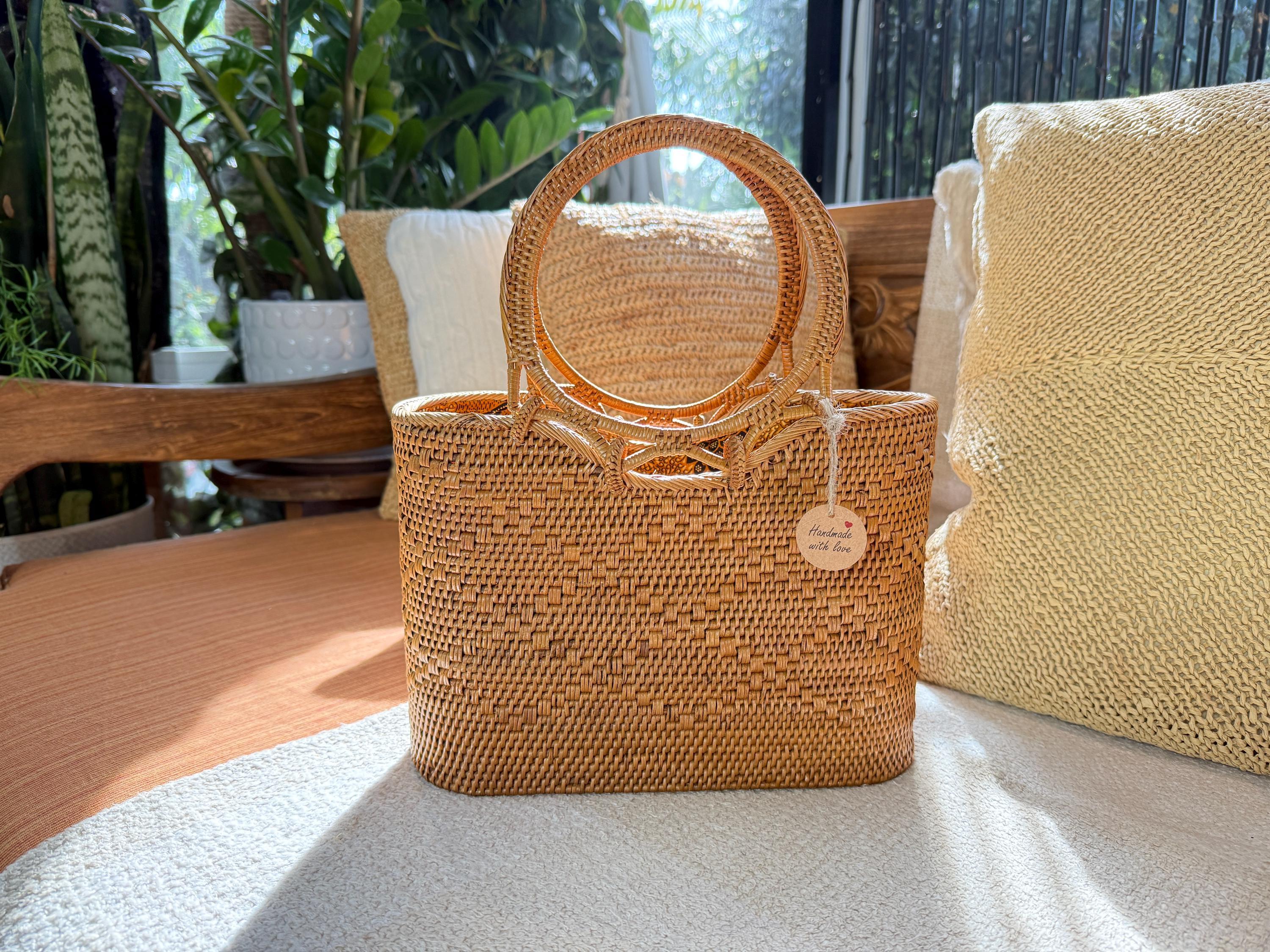 A woven straw handbag with a round handle is placed on a white surface, surrounded by potted plants and a wooden bench.