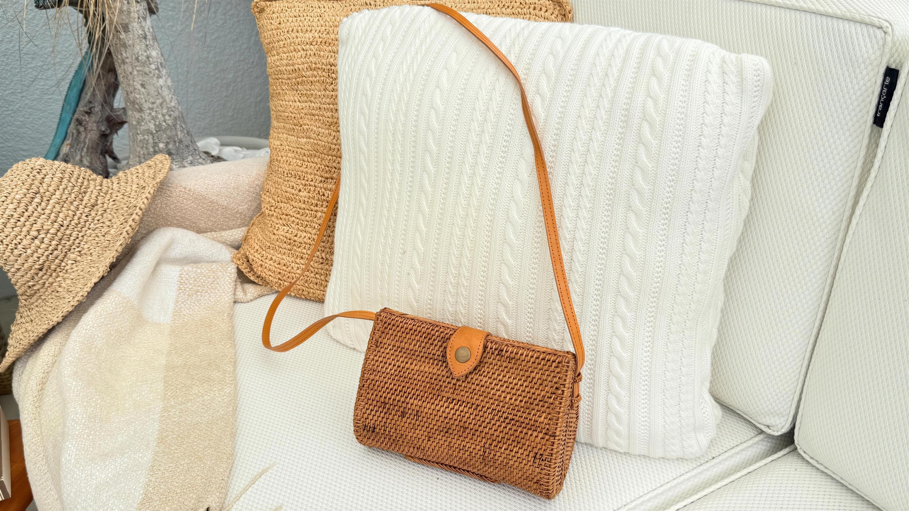 A brown woven purse with a leather strap is placed on a white couch, surrounded by beige and white pillows.