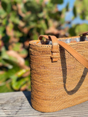 A woven straw bag with a brown leather strap is placed on a wooden surface, with a blurred green background.