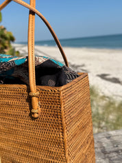 Close-up of the rattan square tote's leather handles and batik drawstring liner at the beach