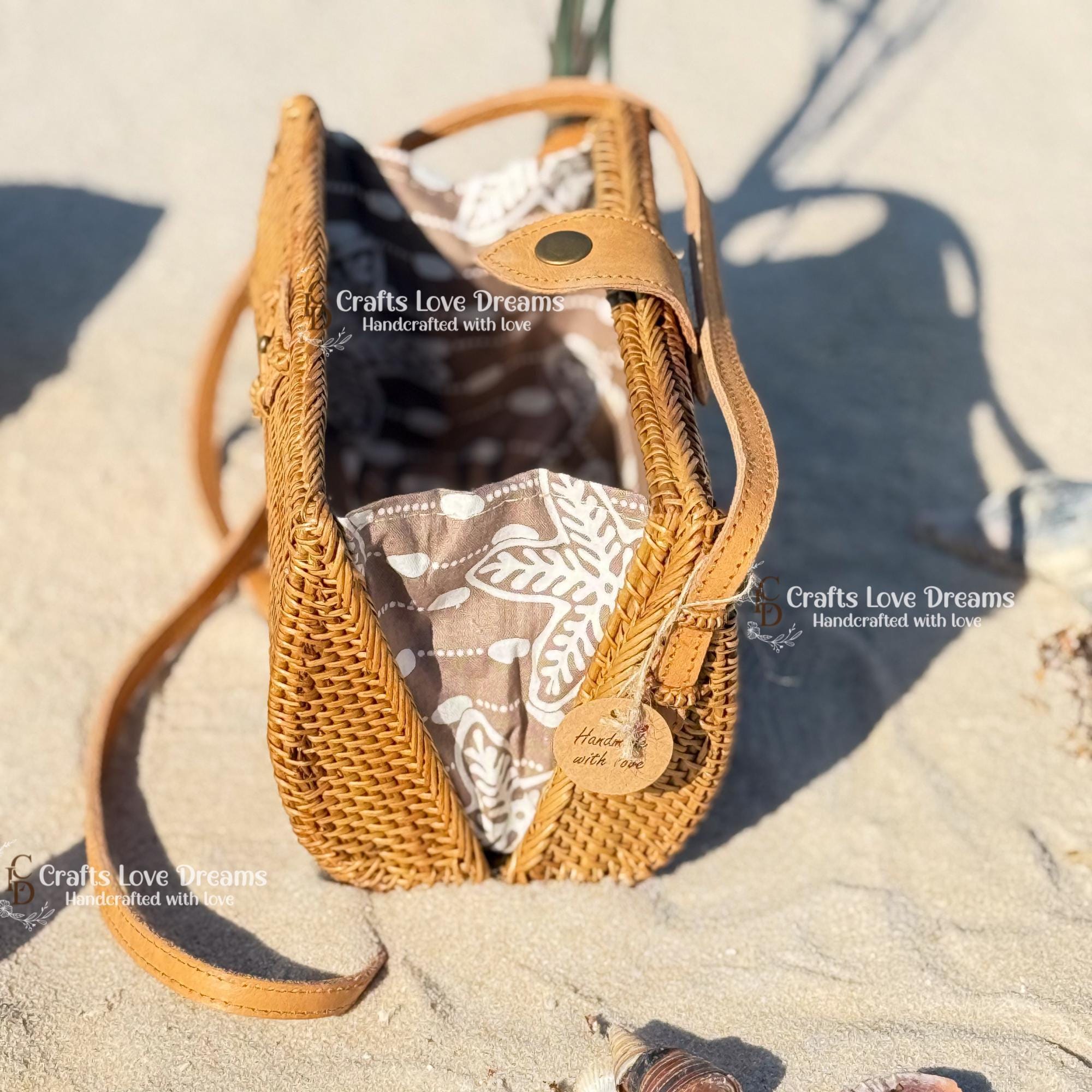 A woven straw handbag with a floral pattern sits on a sandy beach, with the text "Crafts Love Dreams Handcrafted with love" visible.