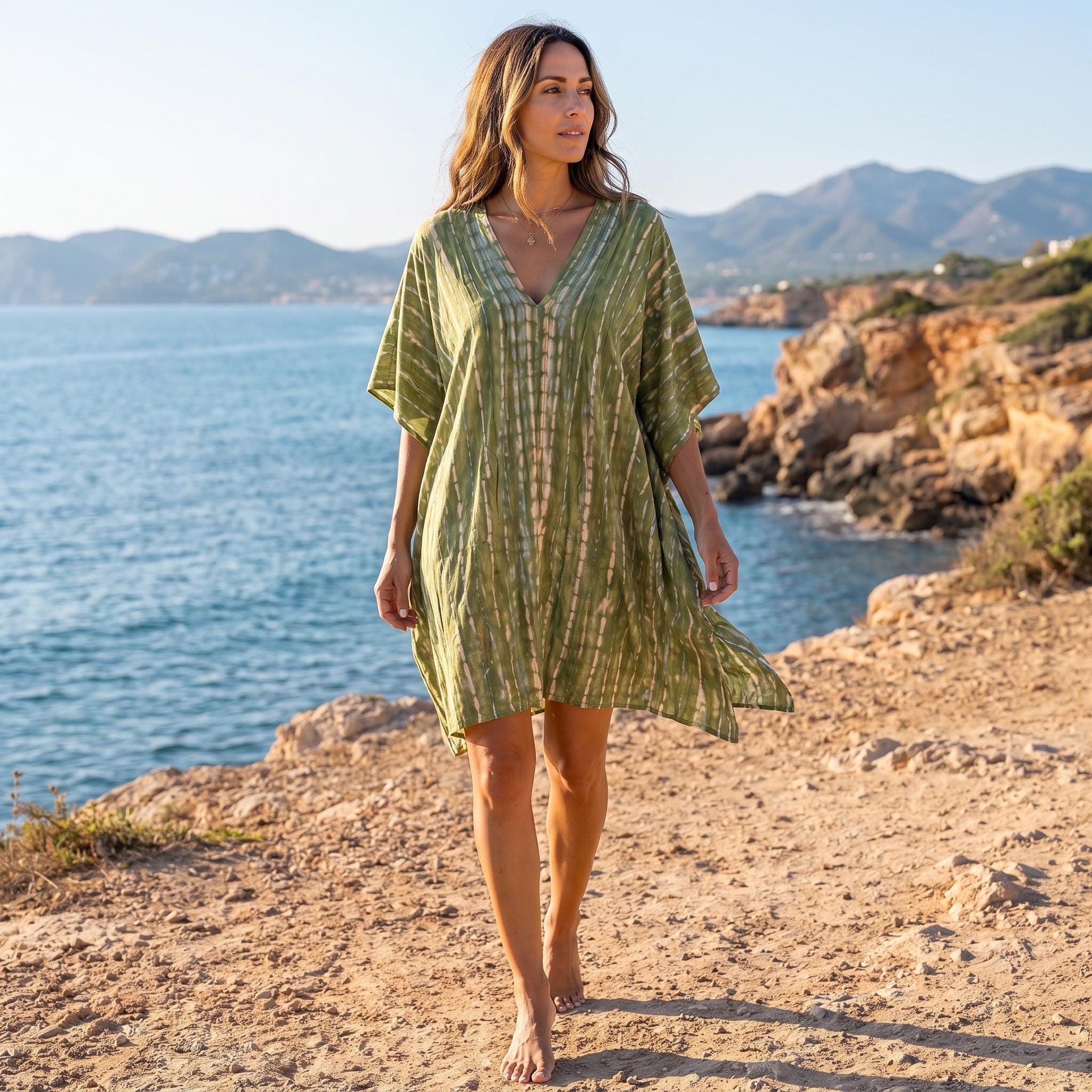 A woman in a green and white striped dress walks along a rocky beach, with mountains visible in the background.