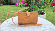 A woven brown purse with a strap is placed on a white table in a garden setting with pink flowers in the background.