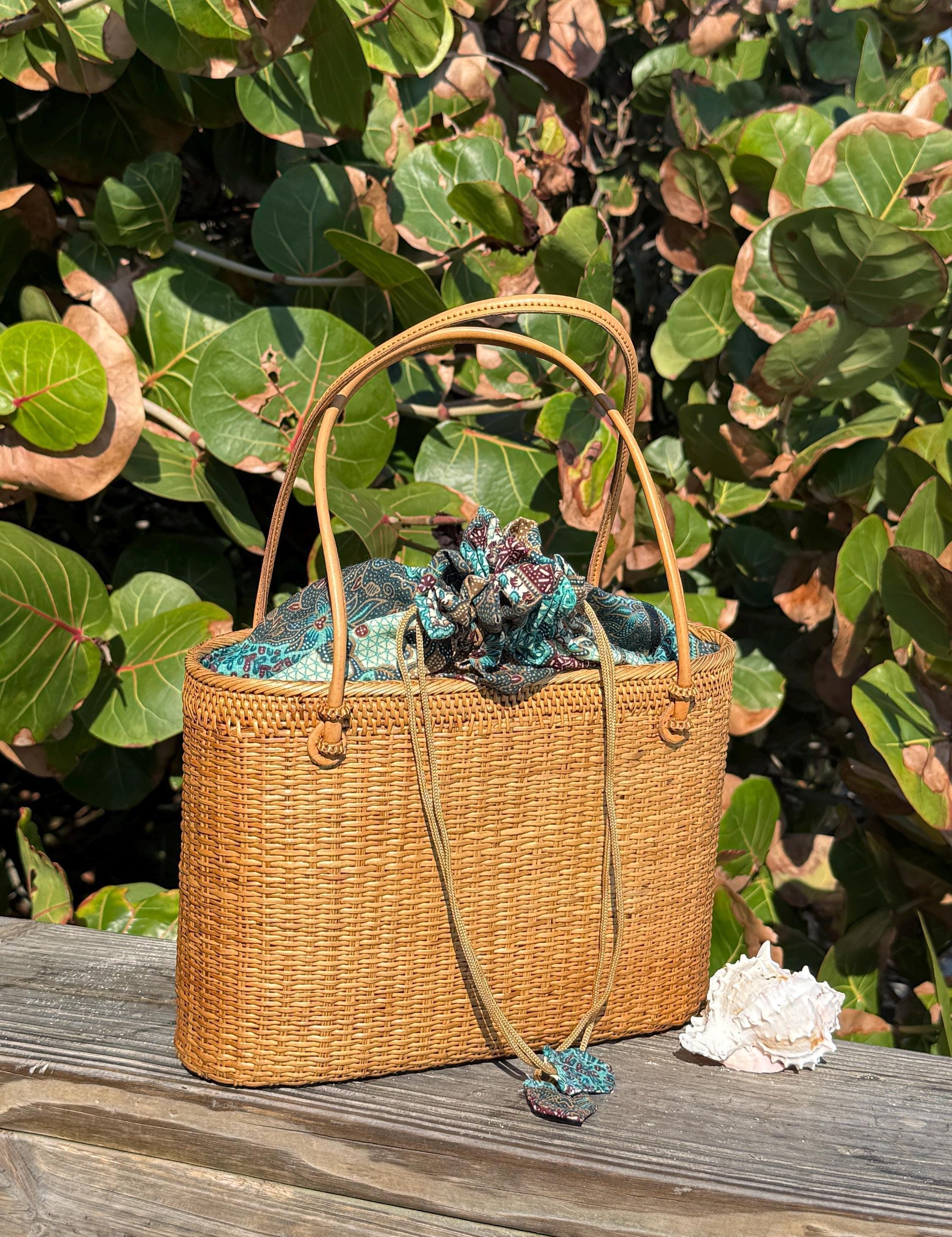 A woven straw handbag with a floral pattern sits on a wooden surface, surrounded by lush green foliage.