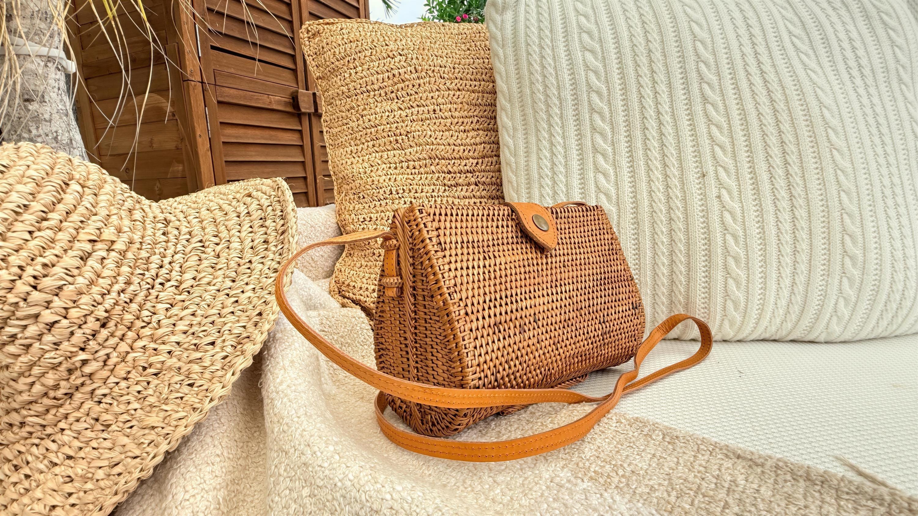 A woven purse rests on a beige blanket, surrounded by straw pillows and a wooden shutter.