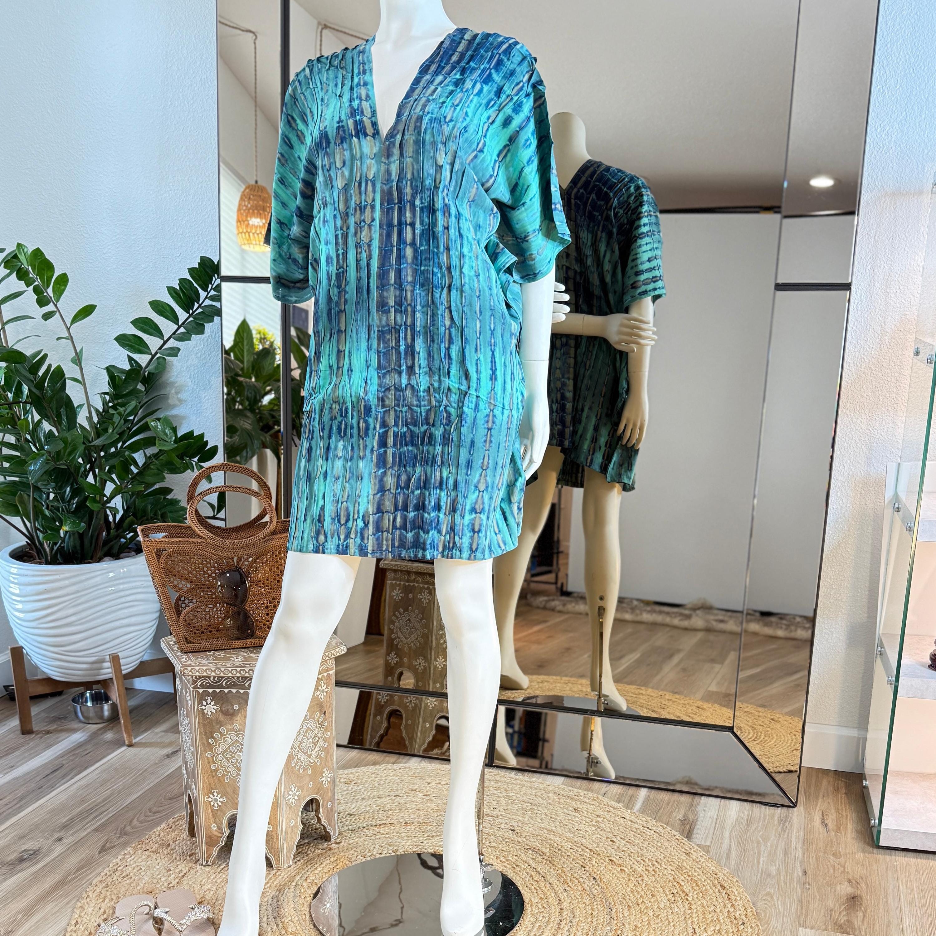 A mannequin wearing a blue and green patterned dress stands in a clothing store, with a mirror and potted plants visible in the background.