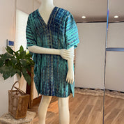 A mannequin wearing a blue and green patterned dress stands in a room with a potted plant and a woven basket nearby.