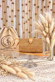 a handcrafted woven purse placed on a glass cake stand, surrounded by decorative elements such as a burlap bag, potted plants, and a vase of dried flowers.