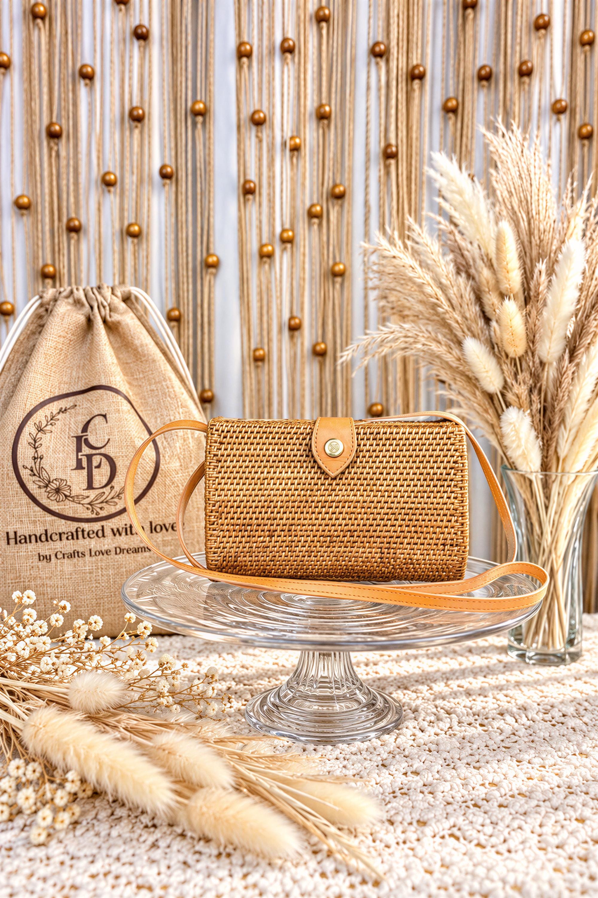 a handcrafted woven purse placed on a glass cake stand, surrounded by decorative elements such as a burlap bag, potted plants, and a vase of dried flowers.