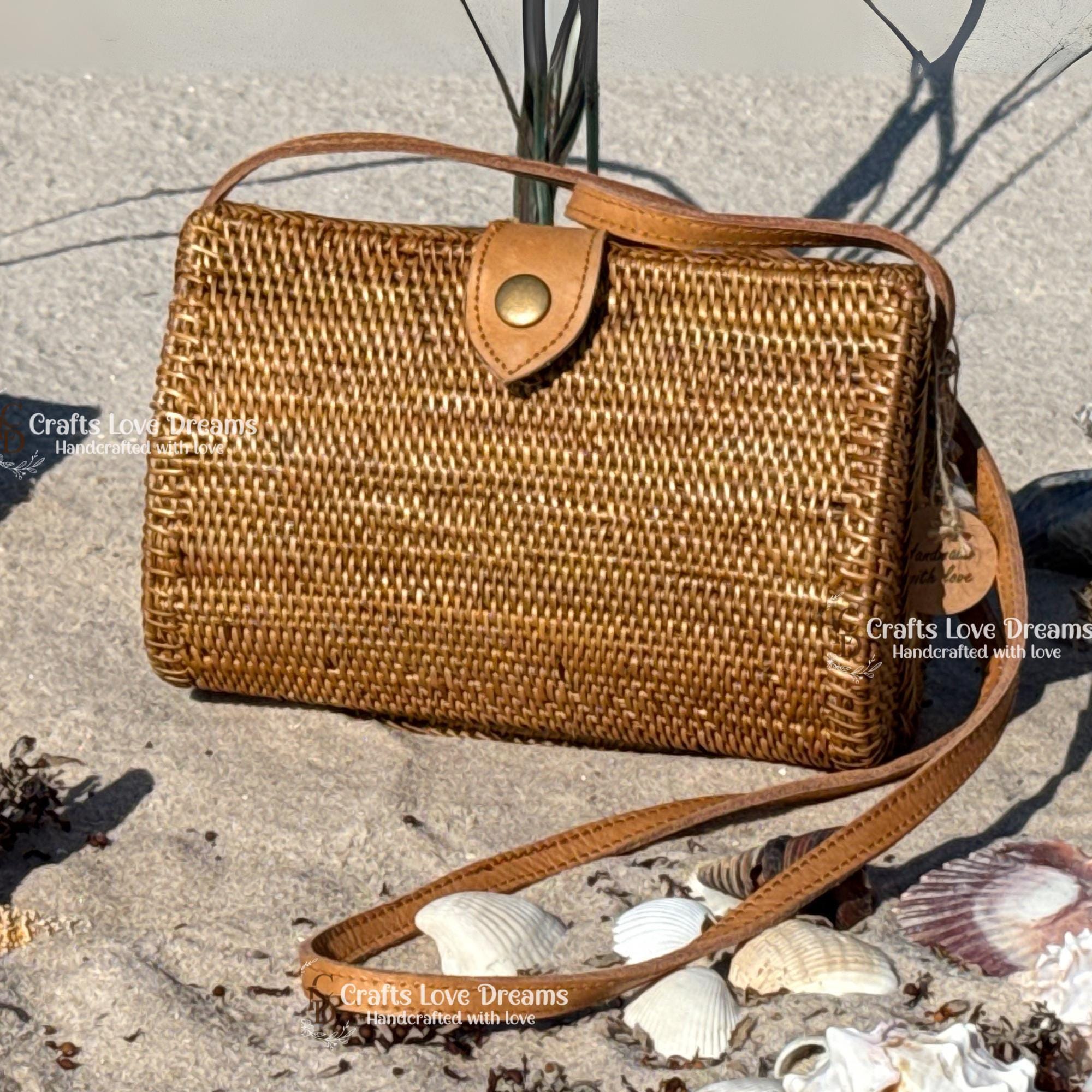 A brown woven handbag with a leather strap sits on a sandy beach, surrounded by seashells.
