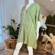 A mannequin wearing a green and white tie-dye dress stands in a clothing store, surrounded by potted plants and other decorative items.