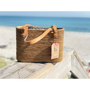 A woven straw handbag sits on a wooden railing, with the ocean visible in the background.