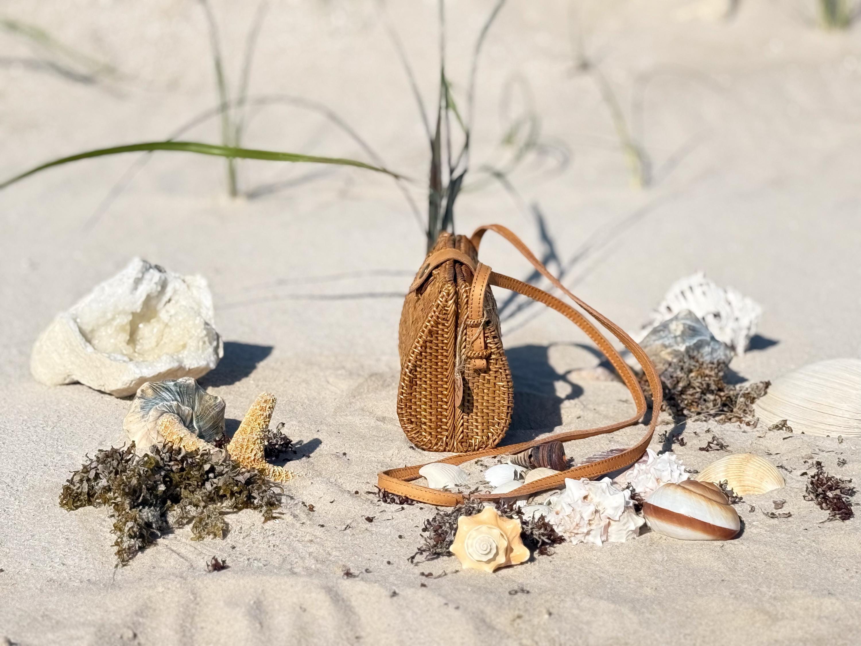 A brown woven purse sits on a sandy beach, surrounded by various seashells and rocks.