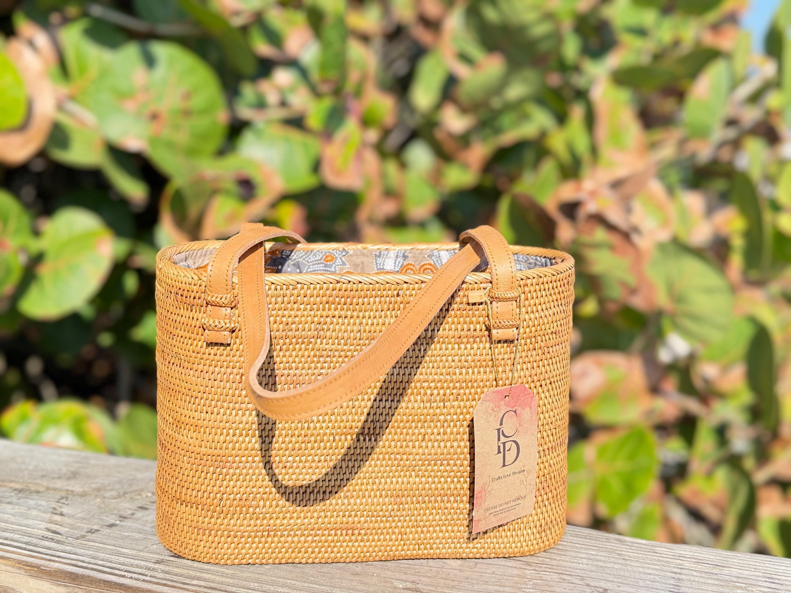 A woven straw handbag with a brown leather strap sits on a wooden surface, with a tag attached to it.