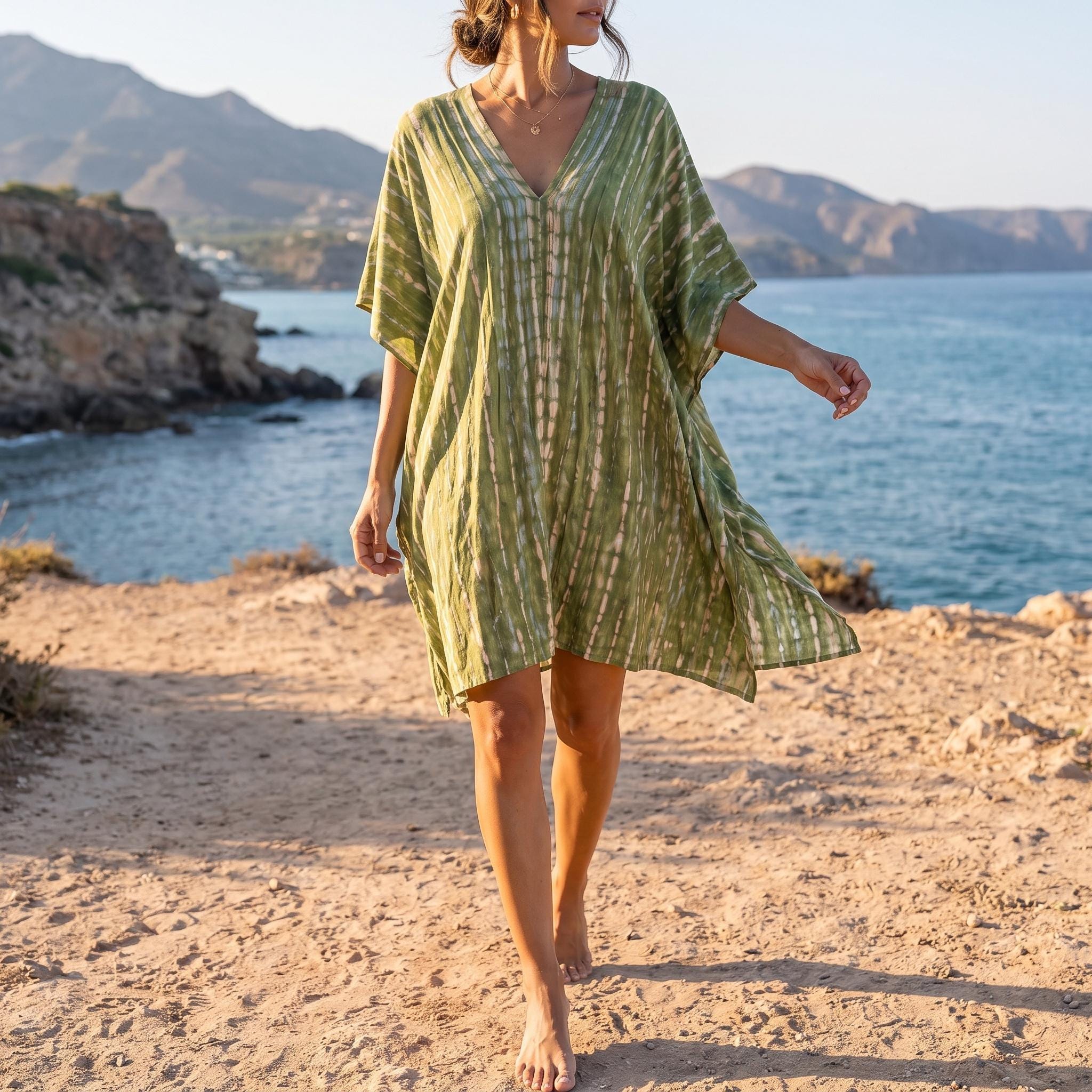 A woman in a green and white striped dress walks along a rocky beach, with mountains and a body of water visible in the background.