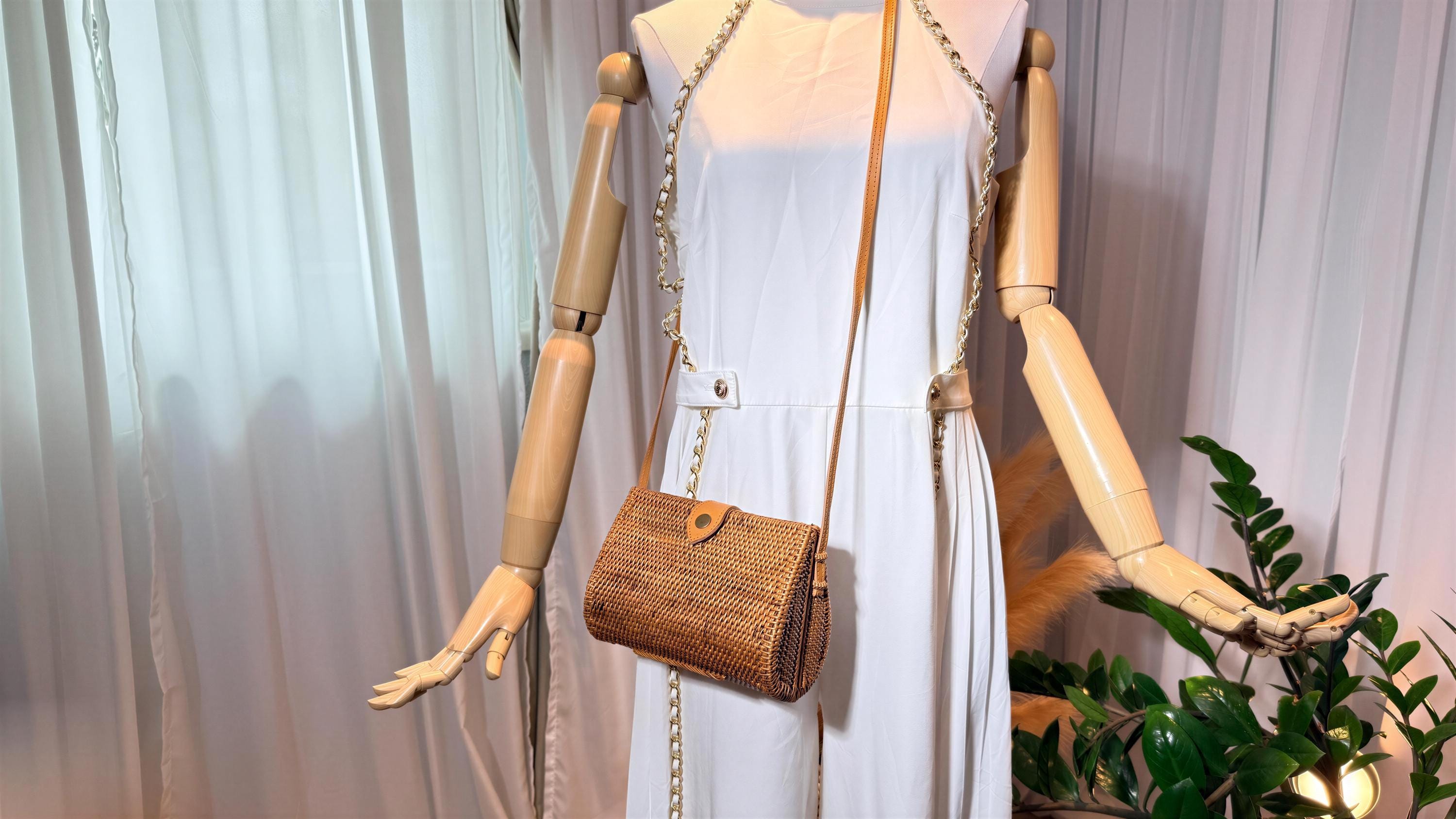 A mannequin wearing a white dress and holding a brown woven purse stands in front of a plant.