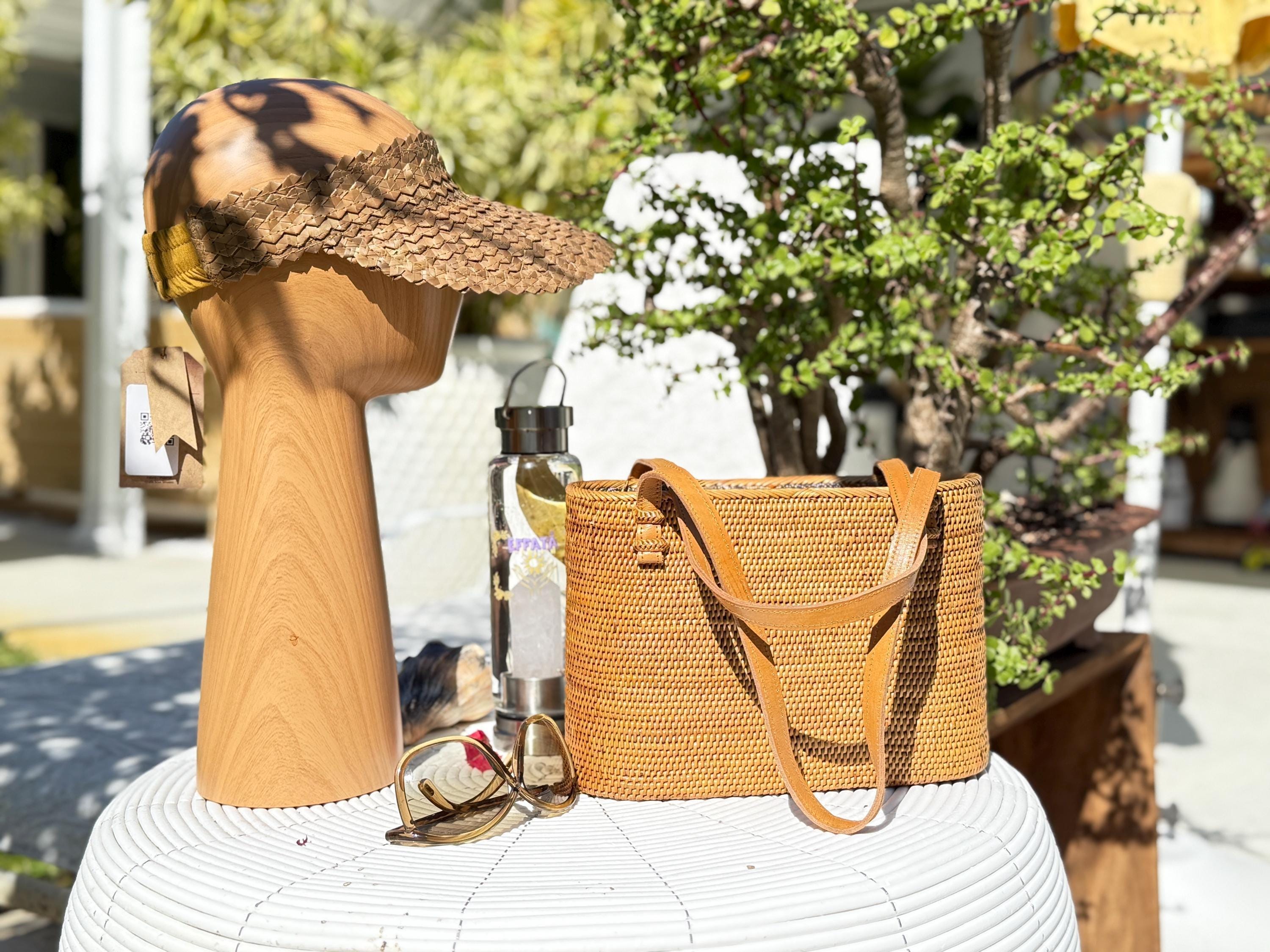 a wooden head with a straw hat, a woven straw bag, a pair of sunglasses, and a glass bottle on a white table.