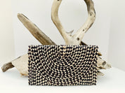 A black and white woven purse with a geometric pattern is displayed against a white background, with a natural branch and driftwood sculpture in the background.