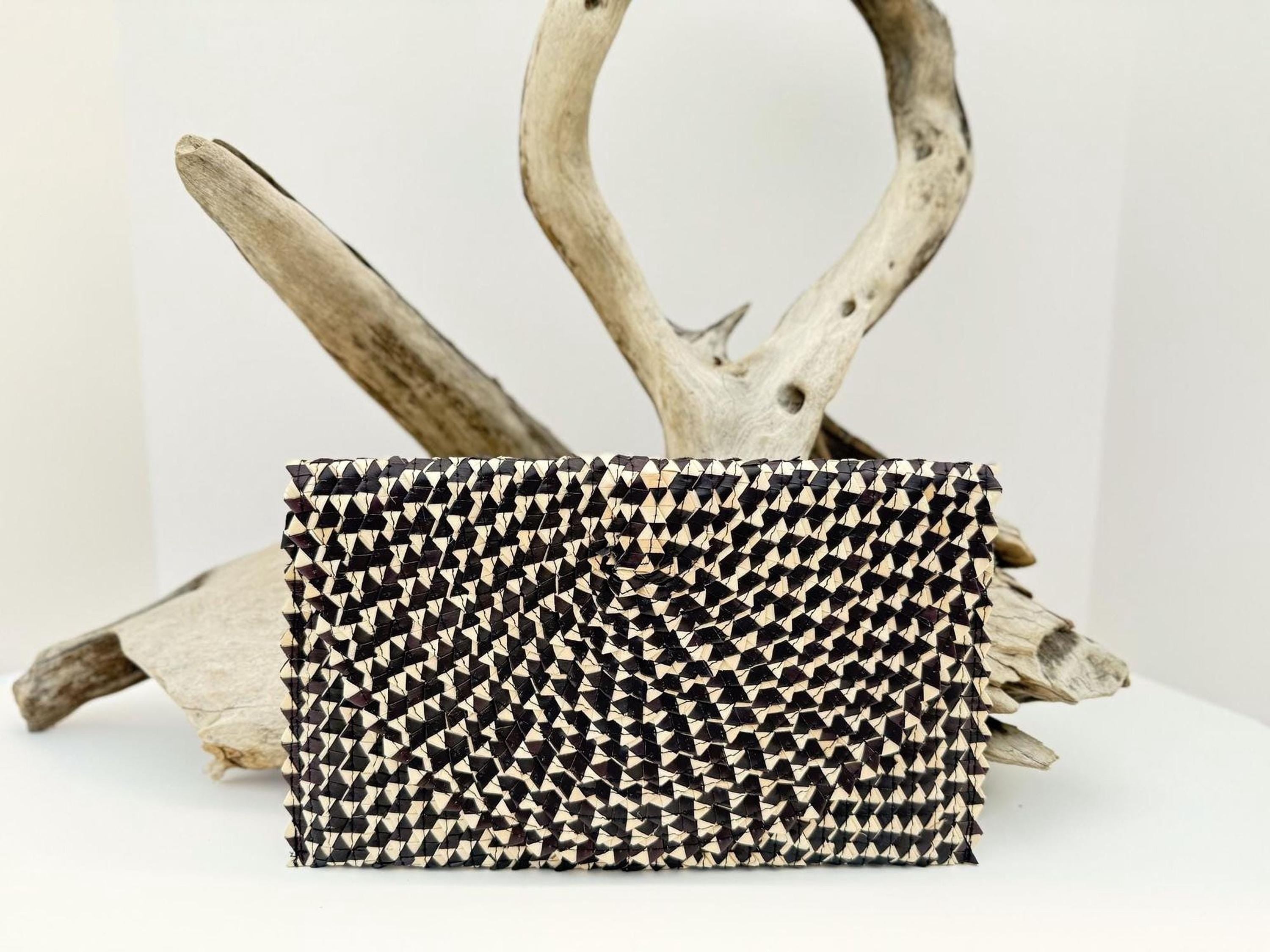 A black and white woven purse with a geometric pattern is displayed against a white background, with a natural branch and driftwood sculpture in the background.