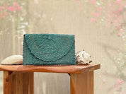 A teal-colored woven purse sits on a wooden table, accompanied by a seashell and a small white object, against a blurred pink and green background.