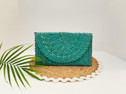 A teal-colored woven purse is displayed on a woven placemat, accompanied by a green leaf.