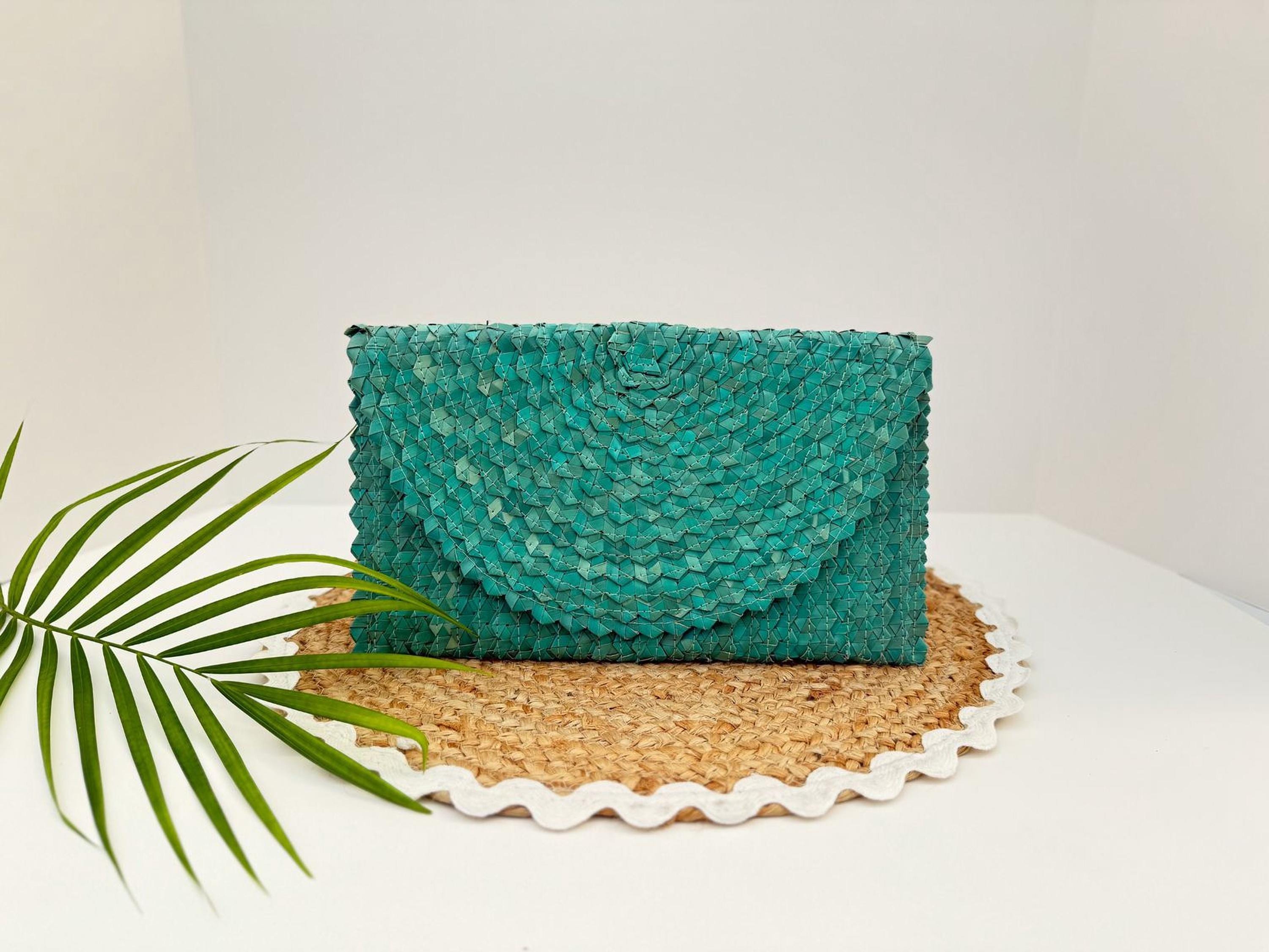 A teal-colored woven purse is displayed on a woven placemat, accompanied by a green leaf.