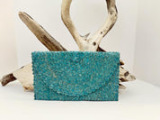 A teal-colored woven purse is displayed against a white background, with a wooden branch and driftwood in the background.