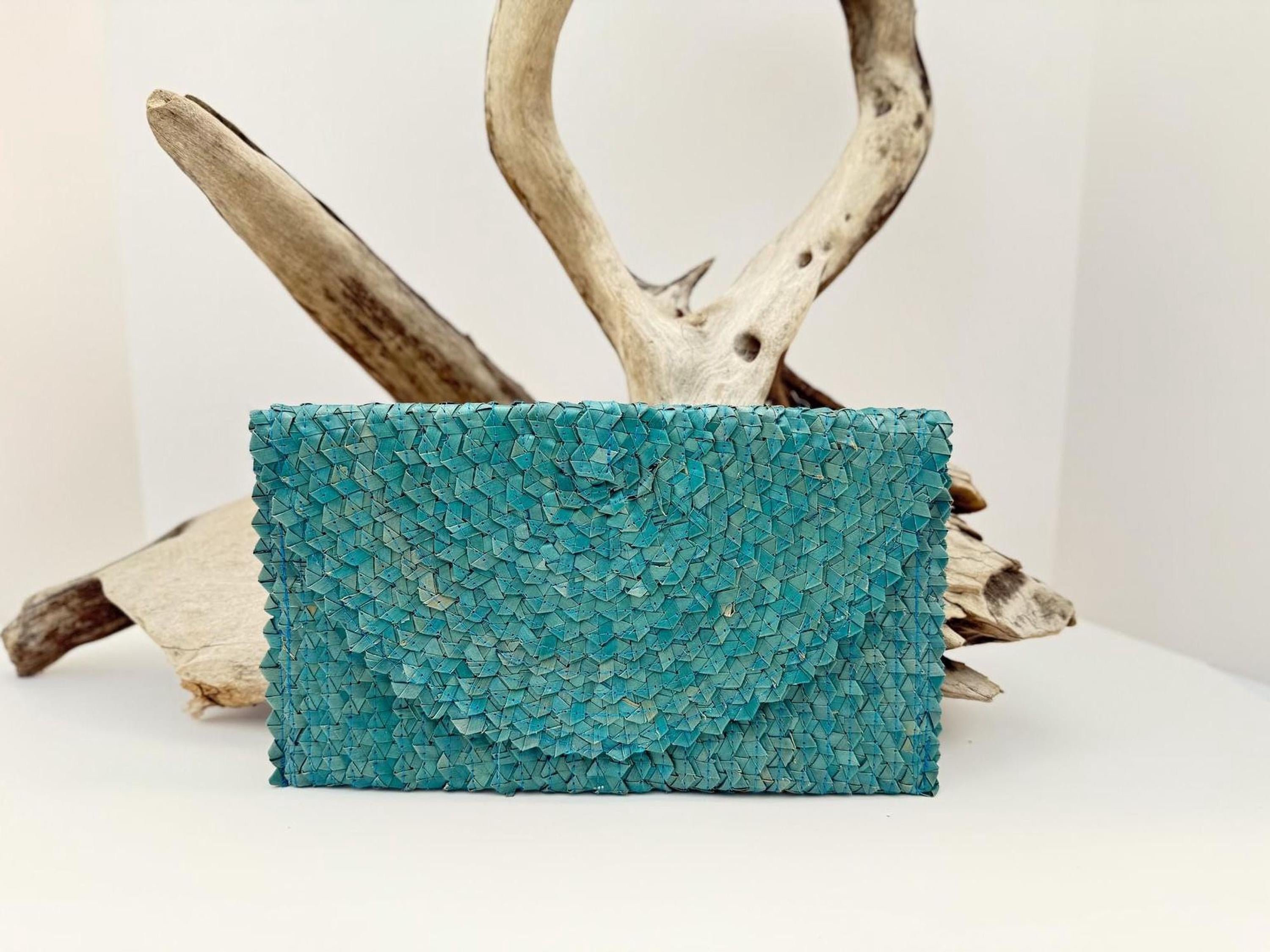 A teal-colored woven purse is displayed against a white background, with a wooden branch and driftwood in the background.