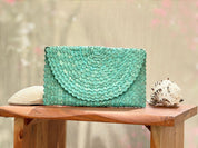 A teal-colored woven purse is displayed on a wooden table, accompanied by a seashell and a small white object.