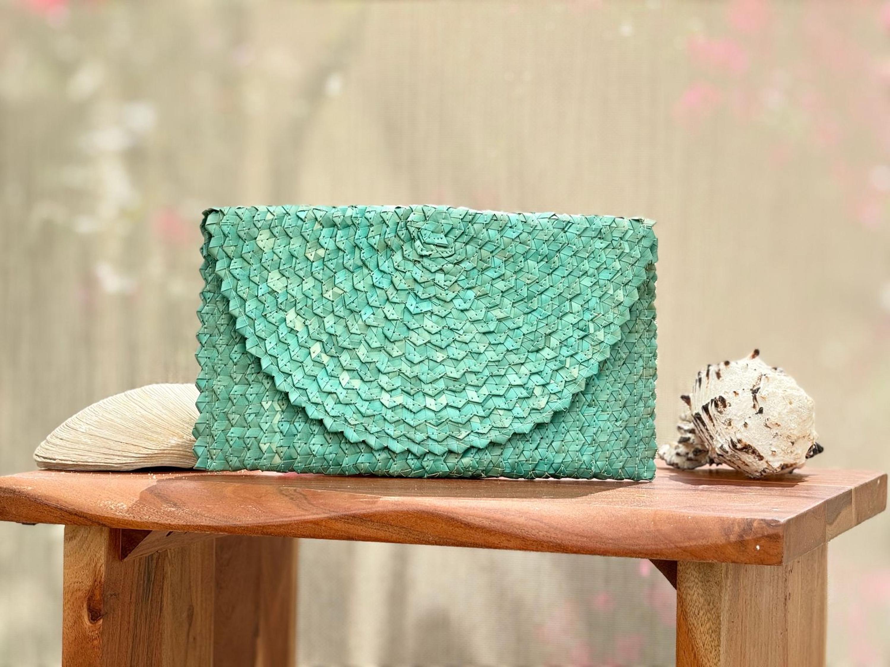 A teal-colored woven purse is displayed on a wooden table, accompanied by a seashell and a small white object.