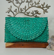 A teal-colored woven purse is displayed on a wooden stand, with a decorative metal tree branch above it.