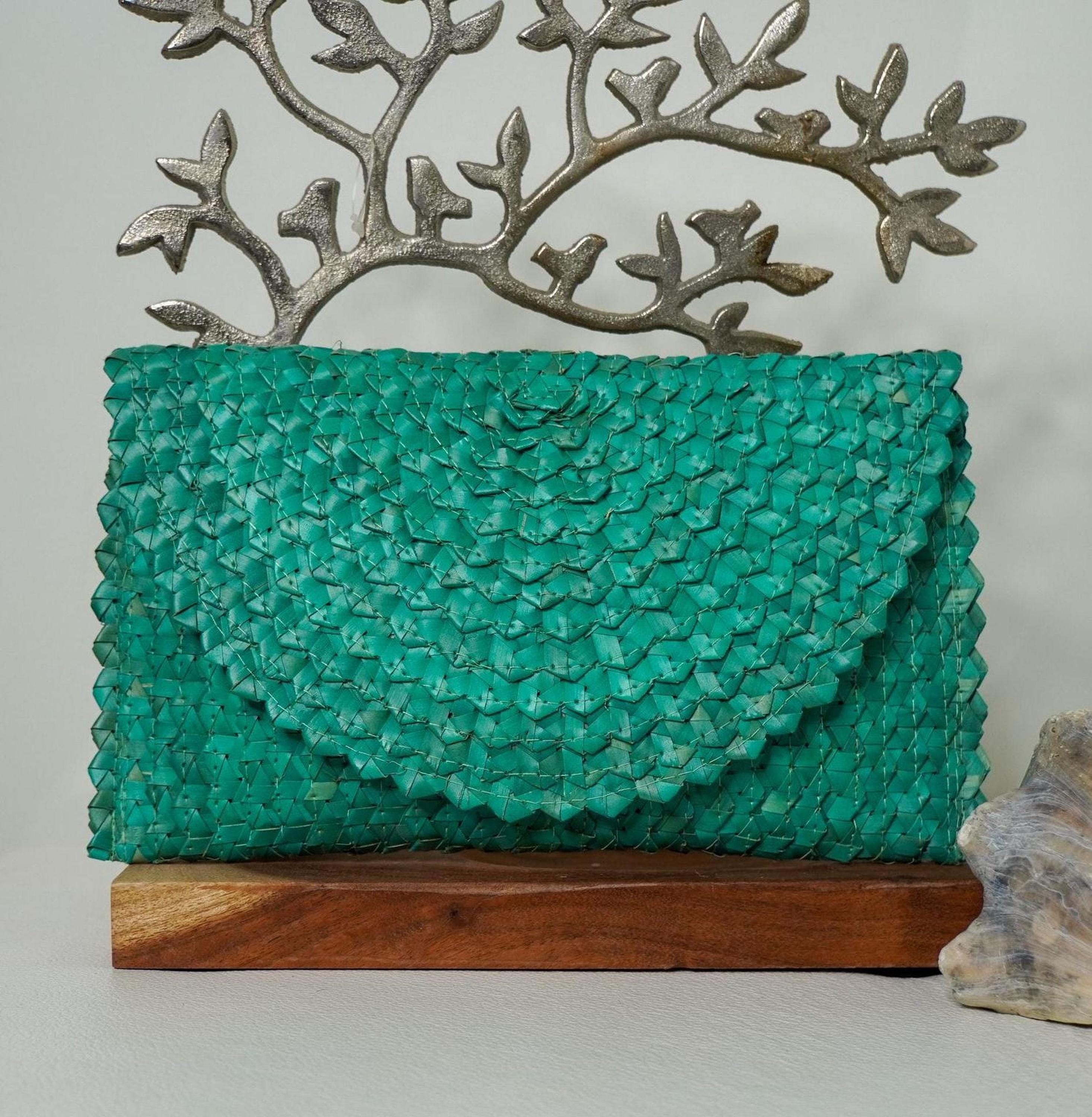 A teal-colored woven purse is displayed on a wooden stand, with a decorative metal tree branch above it.