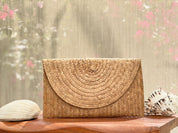 A woven straw purse sits on a wooden surface, with a seashell and a pink flower in the background.