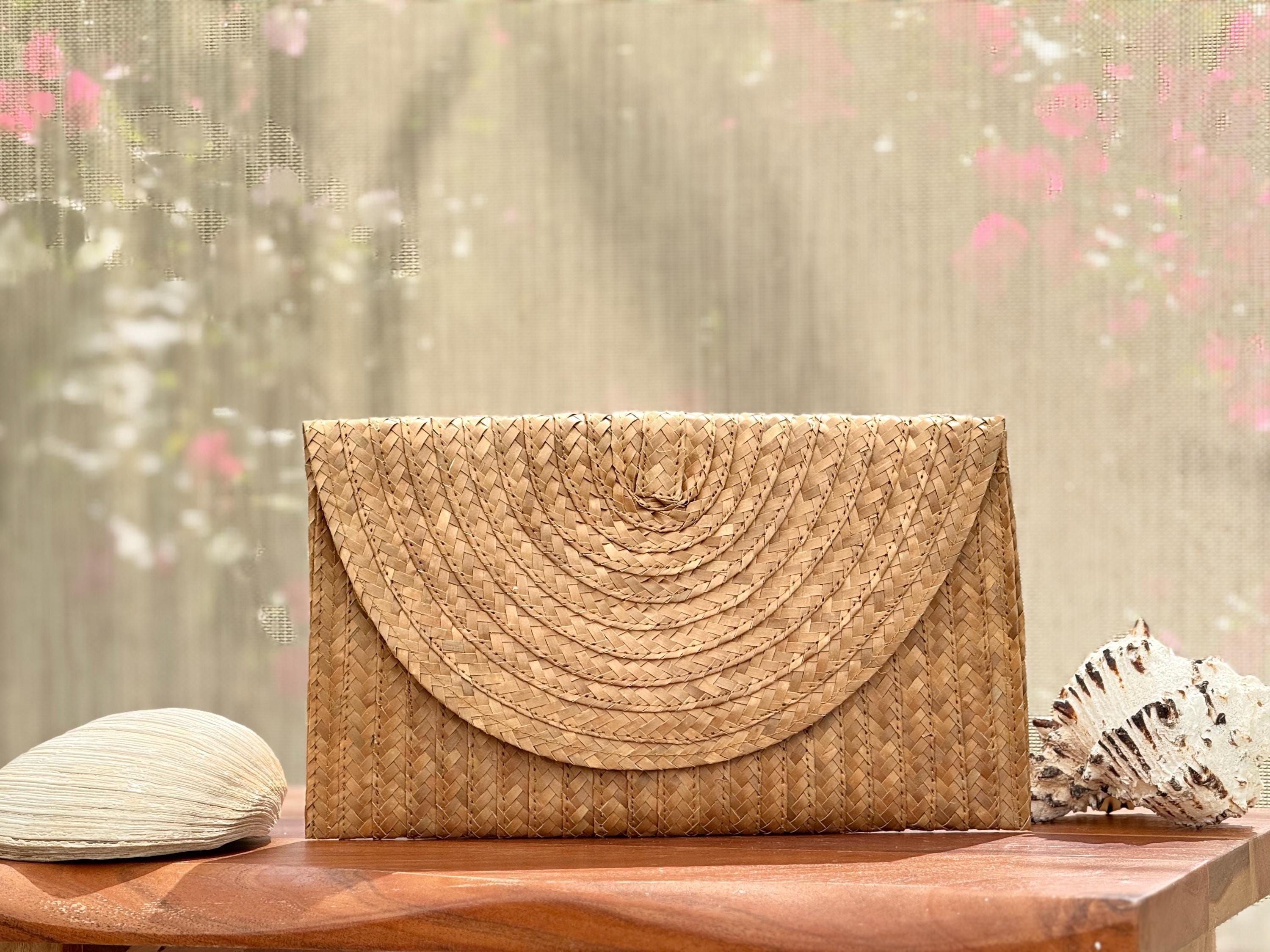 A woven straw purse sits on a wooden surface, with a seashell and a pink flower in the background.