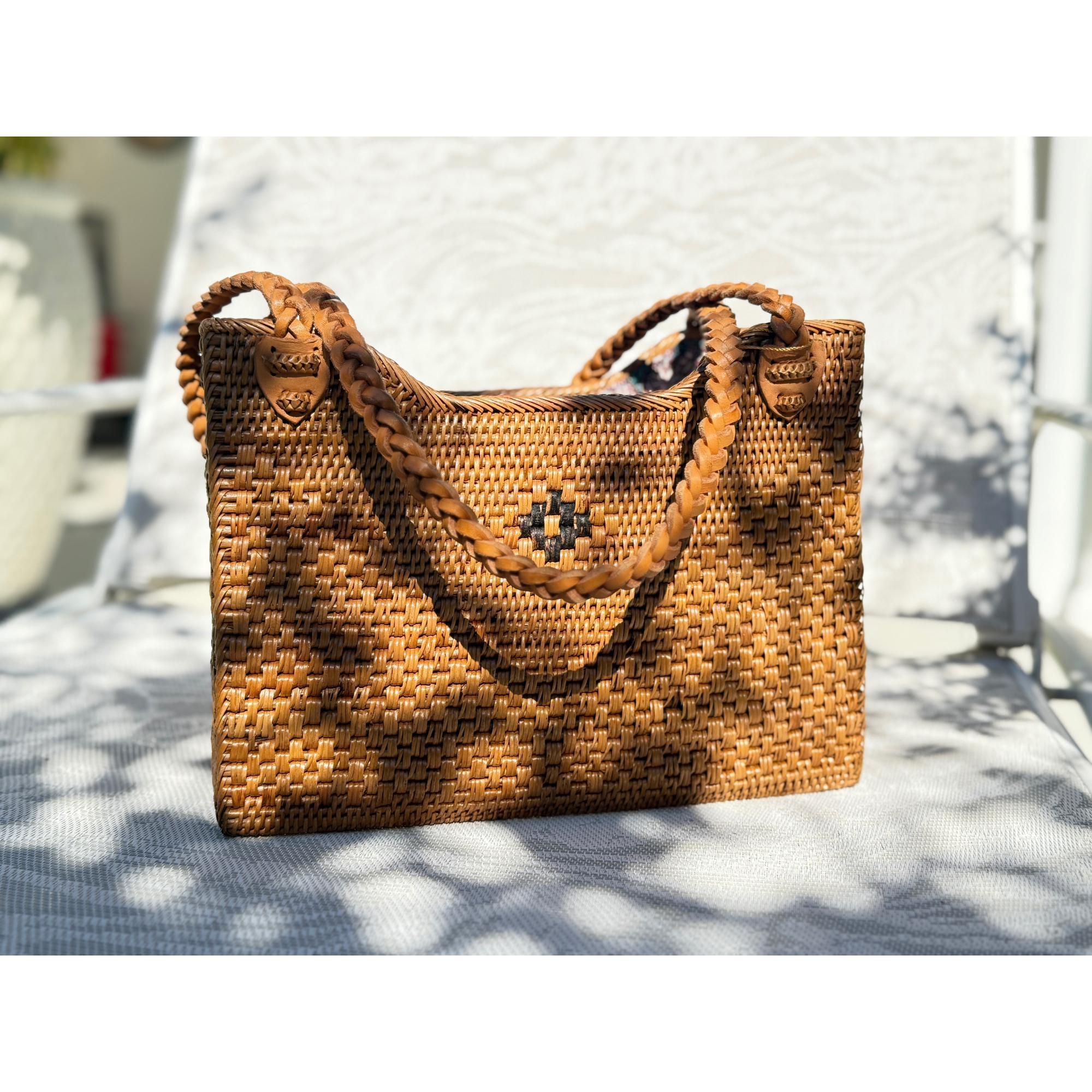 A brown woven handbag with a braided handle is sitting on a white surface, with a blurred background.