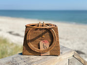 A woven handbag with a round handle sits on a wooden surface, with a tag attached to it, against the backdrop of a sandy beach and the ocean.