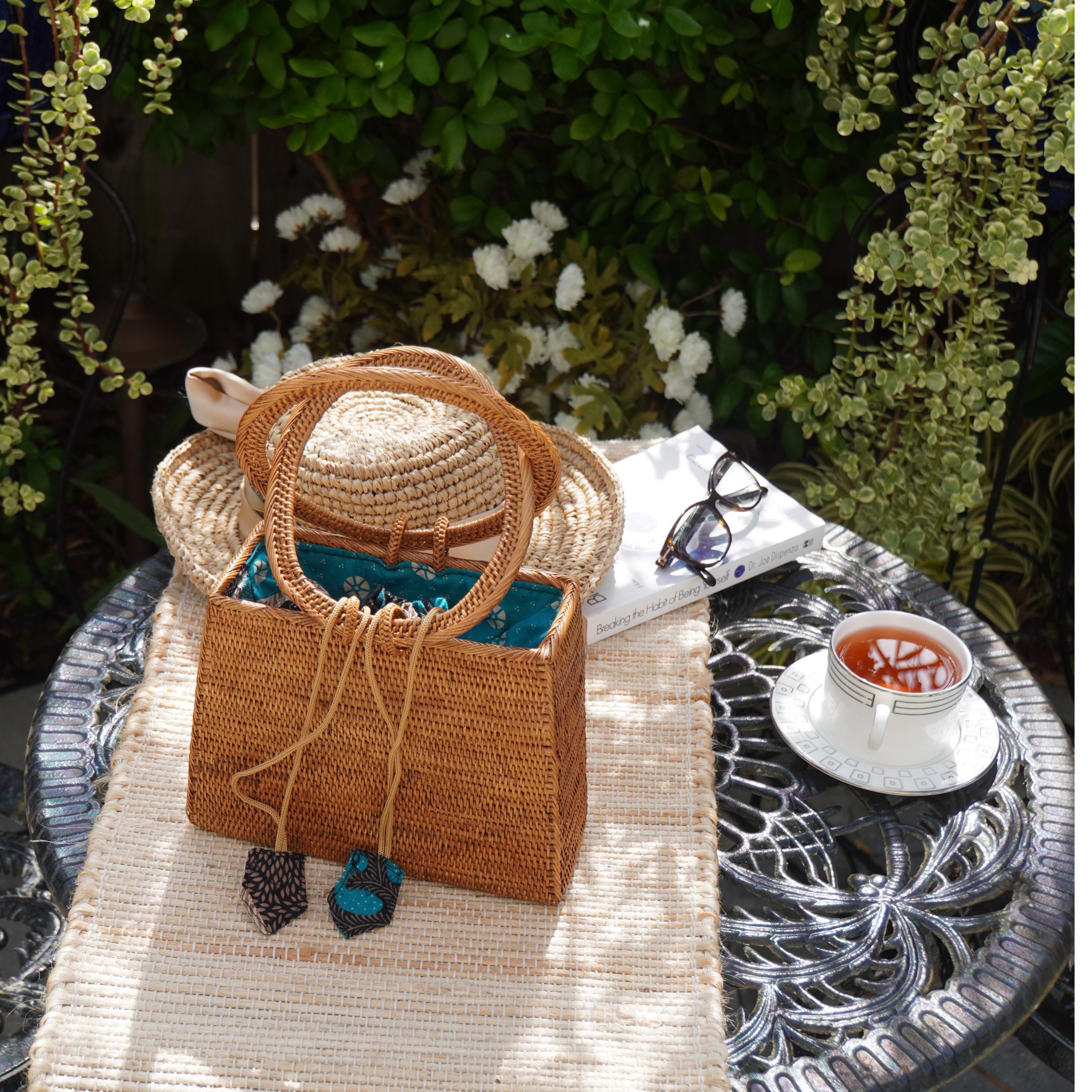 A woven straw bag with a blue and white pattern sits on a beige tablecloth, accompanied by a white book, a white mug with a red beverage, and a pair of sunglasses.