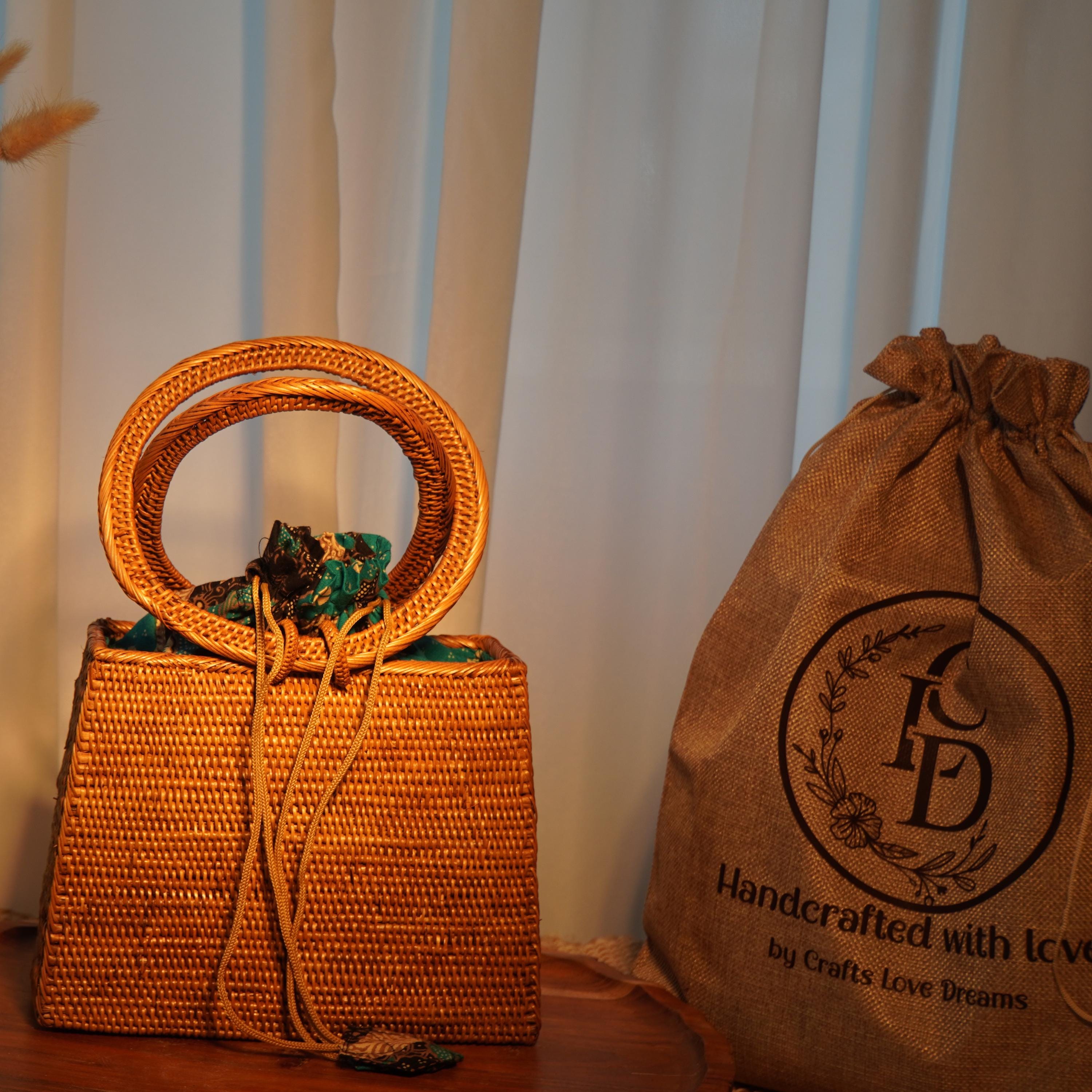 a woven handbag with a round handle and a bag made of burlap with a logo, placed on a wooden surface next to a window with white curtains.