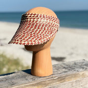 A wooden headless mannequin head wearing a red and white checkered sun visor stands on a wooden surface, with a sandy beach and ocean in the background.