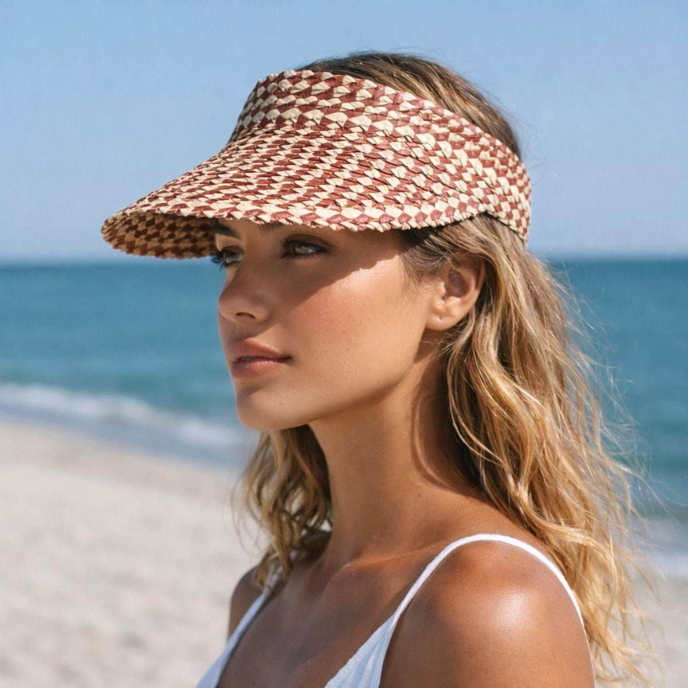 A woman wearing a red and white patterned visor stands on a beach, gazing out at the ocean.