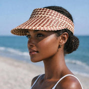 A woman wearing a sun visor and a white tank top stands on a beach, gazing out at the ocean.