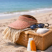 A wicker basket filled with beach essentials, including a red and white sun visor, sunglasses, and a bottle of sunscreen, sits on a sandy beach with the ocean visible in the background.