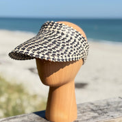 A straw hat with a black and white checkered pattern is displayed on a wooden headless mannequin head, with a blurred beach and ocean in the background.