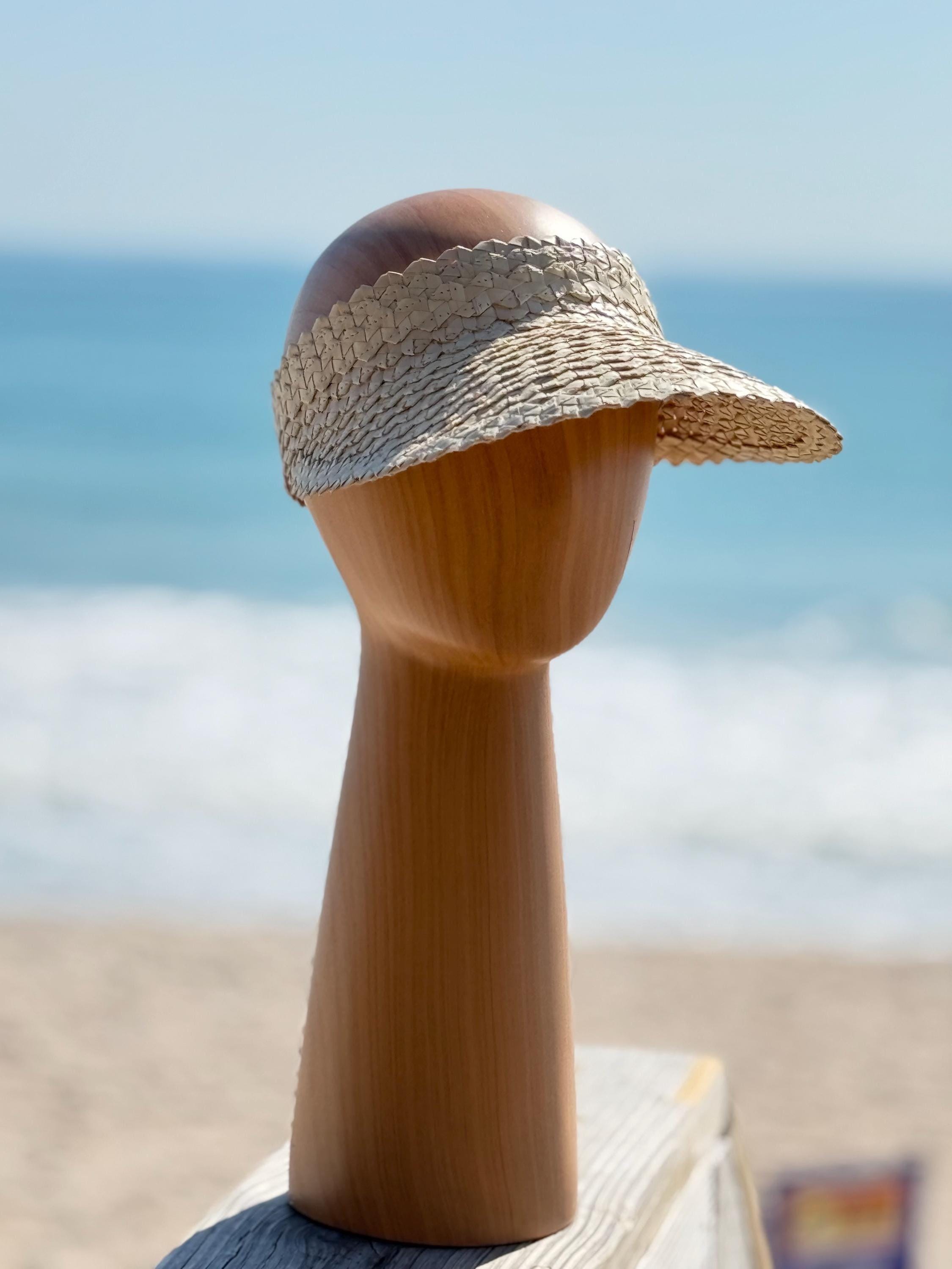 A wooden headless mannequin head wearing a straw hat stands on a wooden post against a backdrop of the ocean.